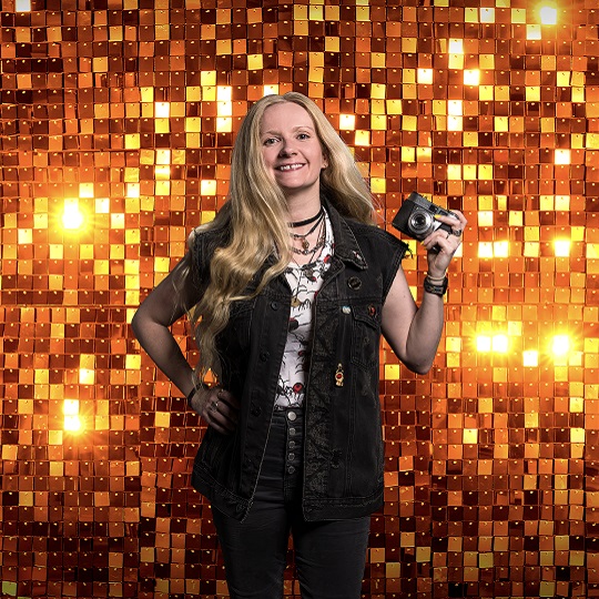  Image of Rhiannon Hopley smiling and holding a camera up in left hand in front of a gold sequined tiled background glinting in the light, Rhiannon has long blond hair and is wearing a sleevless black denim jacket, black jeans and white vest with a black and red print